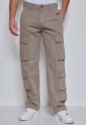 Homme portant un pantalon cargo beige avec plusieurs poches latérales et des baskets blanches, debout devant un fond clair uni.