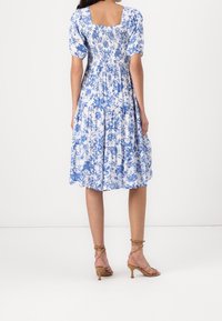 Robe imprimée à fleurs bleues et blanches avec des manches bouffantes, un dos carré, un corsage froncé et une jupe à volants. Associée à des talons beiges à lanières.