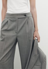 Grijze, geruite, op maat gemaakte broeken met een gladde voorkant, zijzakken en een enkele knoopsluiting, gecombineerd met een bijpassende grijze blazer die in één hand wordt vastgehouden.