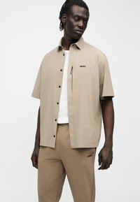 Homme portant une chemise beige à manches courtes boutonnée et un pantalon assorti, tous deux avec le logo "BOSS", debout devant un fond blanc uni.