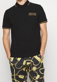 Svart polo skjorta med en gyllene broderad "VERSACE-JEANS COUTURE" logotyp på vänster bröst, med en krage och ärmar med gula kanter.