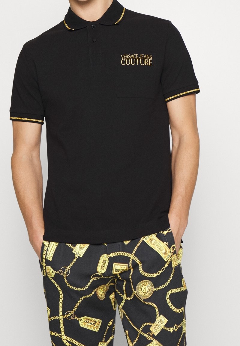Svart polo skjorta med en gyllene broderad "VERSACE-JEANS COUTURE" logotyp på vänster bröst, med en krage och ärmar med gula kanter.