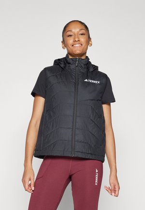 Black padded vest with a high collar, full zipper, and hood; features a logo on the chest. Worn over a short-sleeve black shirt and red leggings.
