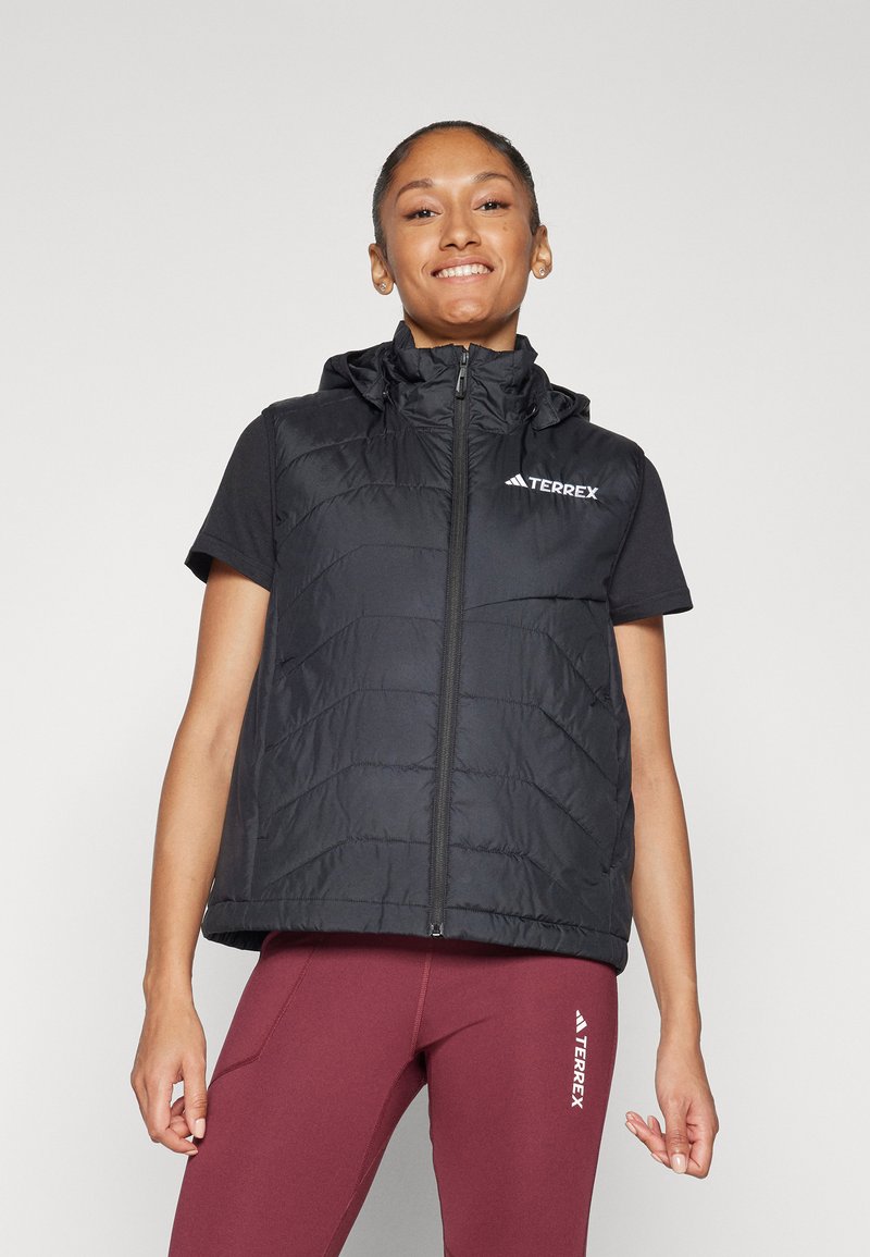 Svart vadderad väst med hög krage, hel dragkedja och huva; har en logotyp på bröstet. Buren över en svart t-shirt med kort ärm och röda leggings.