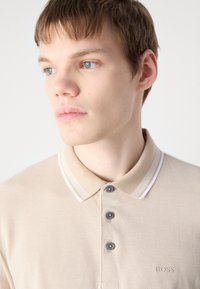 Beige polo krekls ar teksturētu audumu, trim pelēkiem pogām un baltu svītrainu apkakli. Uz krūtīm ir izšūts logo.