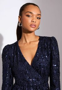 Navy-blaues Paillettenkleid mit tiefem V-Ausschnitt, langen Puffärmeln und taillierter Passform, mit einer strukturierten Oberfläche, die das Licht reflektiert.