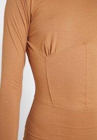 Top bodycon w ciepłym karmelowym kolorze, z długimi rękawami i wyprofilowanym korpusem z marszczeniami na klatce piersiowej. Miękki, elastyczny materiał.