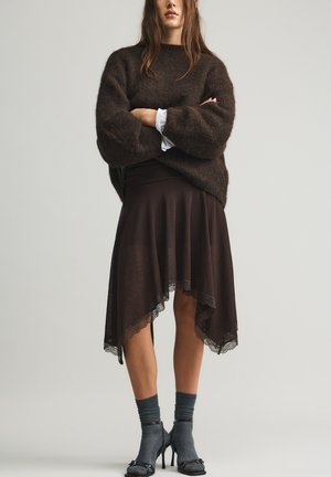 Vrouw draagt een donkerbruine wollige trui, asymmetrische donkere rok met kanten rand, grijze sokken en zwarte hoge hakken, staat met gekruiste armen.