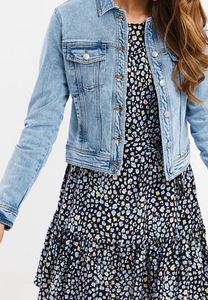 Femme portant une veste en jean bleu clair sur une robe noire avec un petit motif floral coloré, debout devant un fond uni.