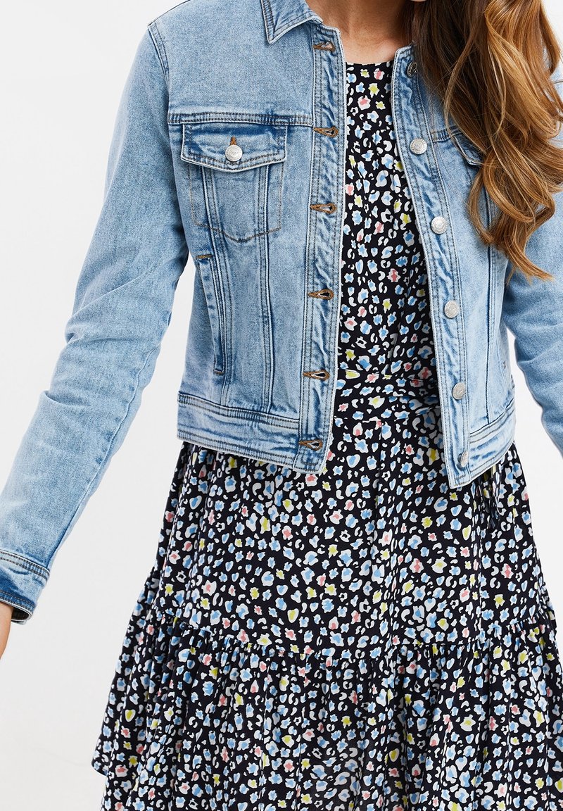 Femme portant une veste en jean bleu clair sur une robe noire avec un petit motif floral coloré, debout devant un fond uni.