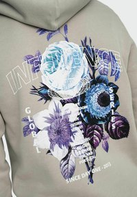 Felpa grigia con ampie grafiche floreali in blu e viola, accompagnata da vari elementi di testo; tessuto morbido e vestibilità rilassata.