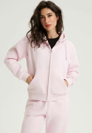 Kobieta z falującymi ciemnymi włosami, ubrana w jasno-różową bluzę z kapturem na zamek oraz dopasowane spodnie dresowe, ręce w kieszeniach bluzy, neutralne tło.
