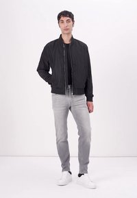 Svart bomberjakke med glidelås foran, ribbede mansjetter og kant, båret over en svart skjorte, sammen med lysegrå jeans og hvite sneakers.