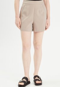 Pantaloni corti beige su misura realizzati in un tessuto morbido, con vita aderente, pieghe e tasche laterali. Abbinati a sandali neri decorati con pois bianchi.