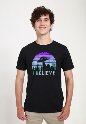Smilende ung mand i sort T-shirt med et farverigt ufo, træer, bjerge og ordene “I BELIEVE”, stående mod en ensfarvet baggrund.