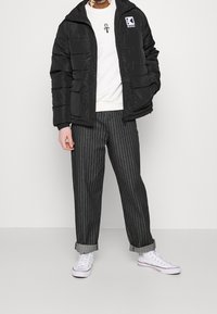 Persona che indossa un giubbotto invernale nero, una felpa bianca con grafica, pantaloni larghi grigi a righe e sneakers bianche alte.