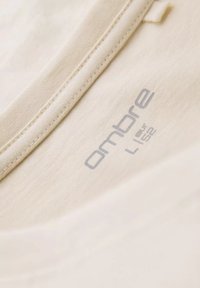 Beige t-shirt stof med en blød tekstur. Har en rund halsudskæring, syet kant, og et gråt "ombre" mærke i kraveområdet. Størrelse L.