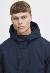 Navy blue jacket with a high collar and oversized hood, featuring a smooth texture and angular seams, designed for a modern, minimal look.