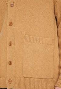 Cárdigan color camello hecho de tela de punto texturizada, con cuello en V, seis botones marrones y un bolsillo parche delantero.