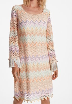 Femme portant une robe en maille à motif zigzag pastel, manches longues finissant par des franges, ourlet festonné et encolure ronde.