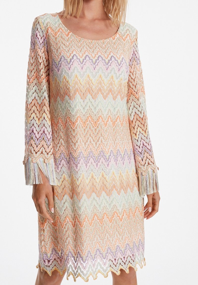 Femme portant une robe en maille à motif zigzag pastel, manches longues finissant par des franges, ourlet festonné et encolure ronde.
