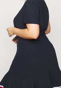 Mujer curvilínea vestida con un vestido azul marino ajustado de mangas cortas y dobladillo acampanado, mostrando un perfil lateral contra un fondo claro.