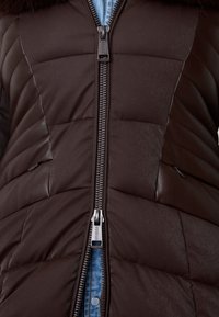 Gilet imbottito marrone con texture trapuntata, dettagli in pelle e zip centrale. Presenta un colletto alto e uno sfondo in denim blu.