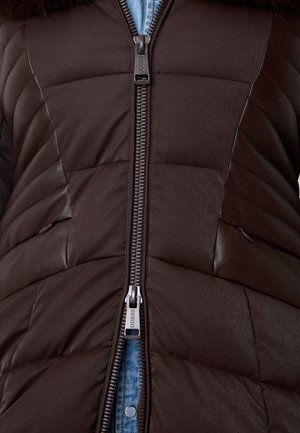 Gilet imbottito marrone con texture trapuntata, dettagli in pelle e zip centrale. Presenta un colletto alto e uno sfondo in denim blu.