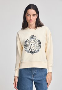 Felpa crema con logo di un club di polo nero e grafica di una corona. Presenta un fit rilassato e maniche lunghe, abbinata a jeans di denim.