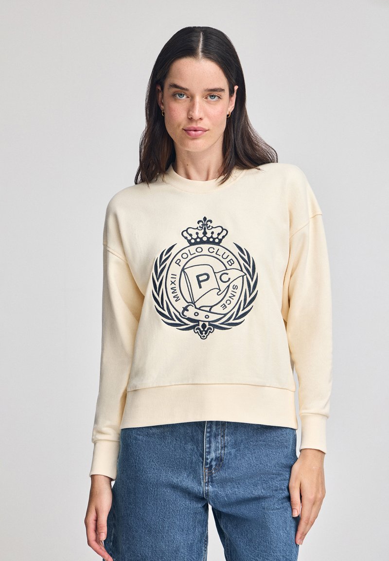 Felpa crema con logo di un club di polo nero e grafica di una corona. Presenta un fit rilassato e maniche lunghe, abbinata a jeans di denim.