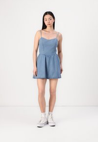 Denim obleka z tankimi naramnicami, prilegajočim bodijem in razširjeno krilo. Svetlo modra barva, neopran rob, oblečena z belimi visokimi superge. Gladka tekstura.