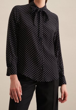 Sort bluse med lange ærmer med hvide polkaprikker, knapper foran og bundet sløjfe ved kraven, båret med sorte bukser.