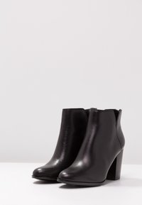 Botas de tobillo de cuero negro con textura brillante, puntera redondeada y tacón cuadrado. Presentan un diseño elegante y minimalista sin hardware visible.