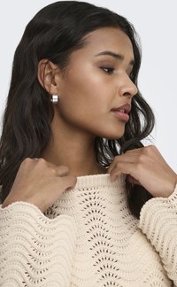 Jeune femme aux cheveux noirs portant des boucles d'oreilles argentées en forme de créoles et un pull en maille crème, ajustant l'encolure avec les deux mains.