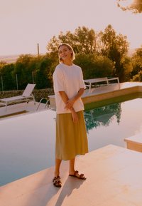 Femme en chemise blanche oversize et jupe olive se tient au bord de la piscine au coucher du soleil, avec des chaises longues et des arbres en arrière-plan.