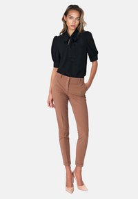 Kobieta w czarnej bluzce z krótkim rękawem i krawatem, w brązowych dopasowanych spodniach i beżowych szpilkach, stojąca z jedną ręką w kieszeni.