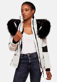 Padded white jacket with black fur hood, zip closure, and ribbed accents. Model wears a white top and dark jeans, showing jacket details.