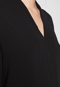 Schwarze Bluse mit V-Ausschnitt, aus glattem Material, das eine minimalistische Ästhetik und klare Linien betont, ohne sichtbare Muster.