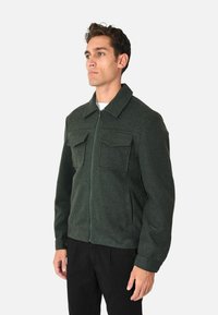 Giacca zip composta in tessuto testurizzato di colore verde scuro, dotata di due tasche sul petto e colletto classico. Indossata con pantaloni neri.