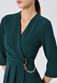 Emeraldgroene wikkeljurk met een diepe V-hals, driekwartmouwen en een decoratieve gouden kettingaccent bij de taille. Gladde stoftextuur.