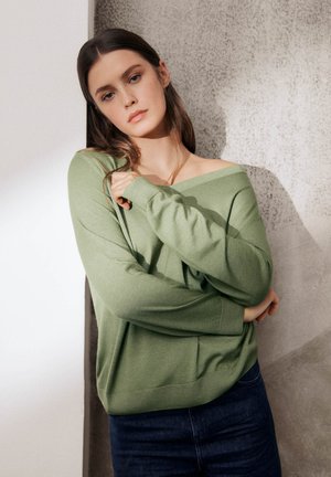 Jeune femme aux longs cheveux bruns, portant un pull vert à épaules dénudées et un jean foncé, debout contre un mur texturé.