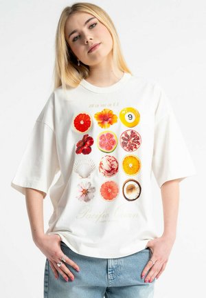 Młoda kobieta o długich blond włosach nosi oversize białą koszulkę z dziewięcioma plasterkami owoców i kwiatów ułożonymi w siatkę, zestawioną z jasnoniebieskimi dżinsami.