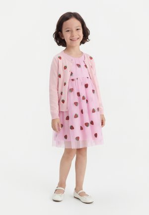 Bambina sorridente che indossa un vestito rosa e un cardigan con motivi di fragole, in piedi con scarpe bianche davanti a uno sfondo semplice.