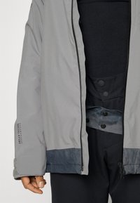 Veste imperméable grise avec fermeture éclair, présentant des accents bleu foncé au bas et aux poignets. Comprend un détail de logo et une couche intérieure boutonnée.