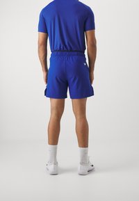 Blå atletiska shorts med elastisk midja, sidofickor och subtil logotypdetalj. Bärs med vita strumpor och skor. Bakifrån.