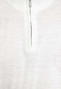 Weißer gestrickter Pullover mit hohem Kragen und kurzem Reißverschluss. Er hat eine weiche Textur und ein dezentes, offenes Strickmuster.