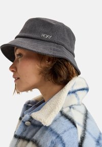 Gray corduroy bucket hat with a stitched "ROXY" logo. Features wide brim and textured fabric. Worn with a blue and white checkered jacket.