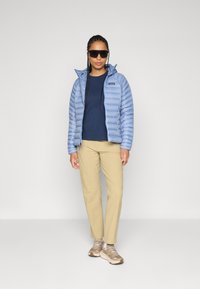 Žena nosi plavu puffer jaknu, tamnoplavu majicu, bež hlače, smeđe sportske tenisice i tamne velike sunčane naočale, dok stoji protiv jednostavne svijetle pozadine.