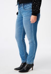 Jean slim bleu clair avec taille élastique et cordon de serrage. Présente des détails de couture et un léger délavage, associés à des bottines noires à talons.