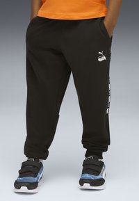 Kind trägt schwarze Puma-Sweatpants mit Hot-Wheels-Logo an der Seite, kombiniert mit blauen und schwarzen Turnschuhen auf grauem Hintergrund.